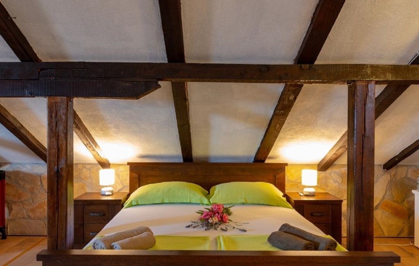 A four-poster bed with a stone wall backdrop, wooden beams, and two bedside lamps.