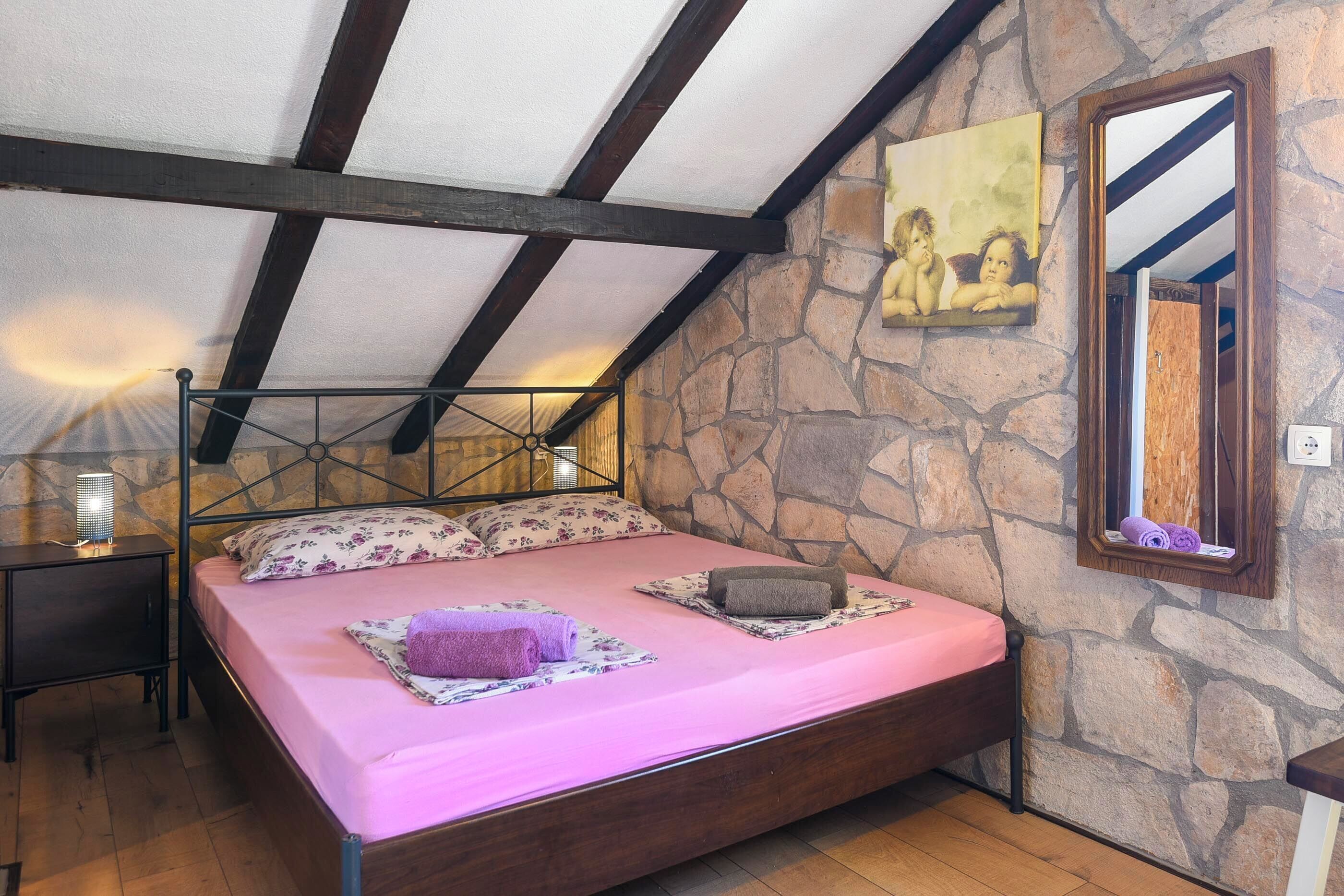 Ein Schlafzimmer mit Steinsturz, einem Bett mit rosa Bettwäsche, einem Holz-Kopfstück, einem Nachttisch, einer Lampe und einem Spiegel.