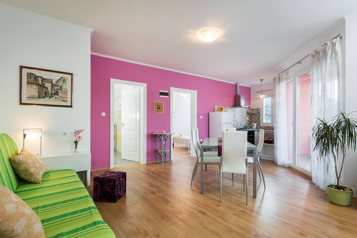 Ein heller Wohnraum mit einer rosafarbenen Wand, einem grünen Sofa, einer Essecke mit Stühlen und einer Küche im Hintergrund.