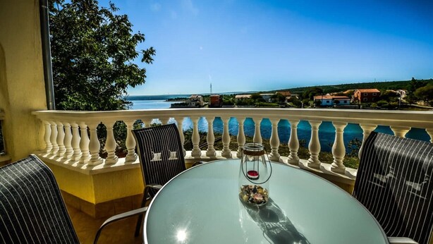 A balcony with a glass table and chairs, overlooking a coastal view with houses and a clear sky. - Apartment Roko (Obrovac)