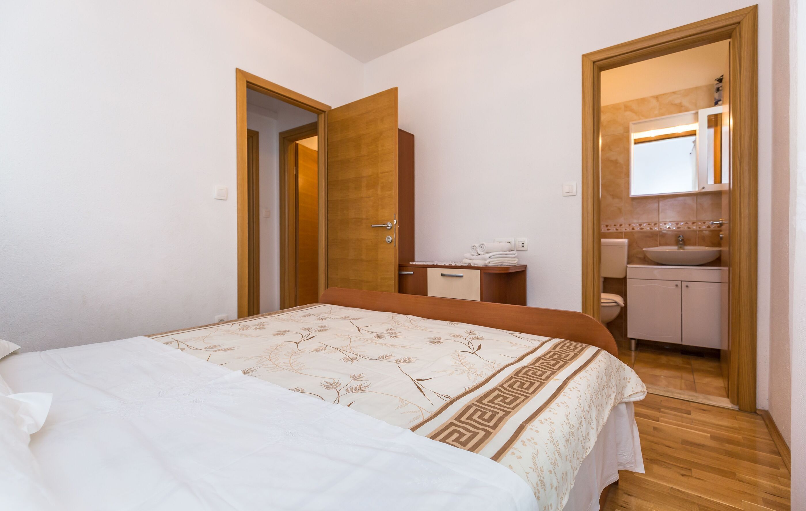 A bedroom with a bed, a wooden door, a small bedside table, and a bathroom with a sink and mirror.