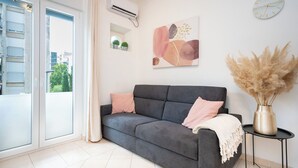 Ένα μοντέρνο σαλόνι με έναν γκρι καναπέ, ένα μικρό βοηθητικό τραπεζάκι, ένα επιτοίχιο ρολόι και ένα παράθυρο με κουρτίνες.