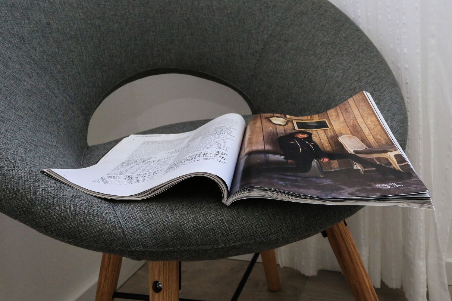 En öppen tidning ligger på en grå klädd fåtölj med ben av trä.