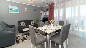 Un salon moderne comprenant un coin repas, des canapés gris, une table en verre et une grande fenêtre ornée de rideaux légers.