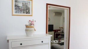 En vit byrå med en inramad tavla ovanför, en vas med blommor på byrån och en spegel som speglar ett rum med en säng och en tv.