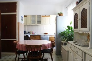 Μια μικρή, λειτουργική κουζίνα  με λευκά ντουλάπια, ένα τραπέζι φαγητού με καρέκλες, ένα ψυγείο και ένα φυτό σε γλάστρα.