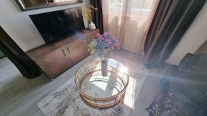 Una sala de estar con un televisor sobre un mueble de madera, una mesa redonda de centro con un jarrón de flores, y vista al exterior a través de amplios ventanales.