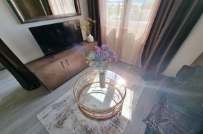 A living room with a television on a wooden cabinet, a round coffee table with a vase of flowers, and a view of the outdoors through large windows. - Apartment Isa (Zadar)