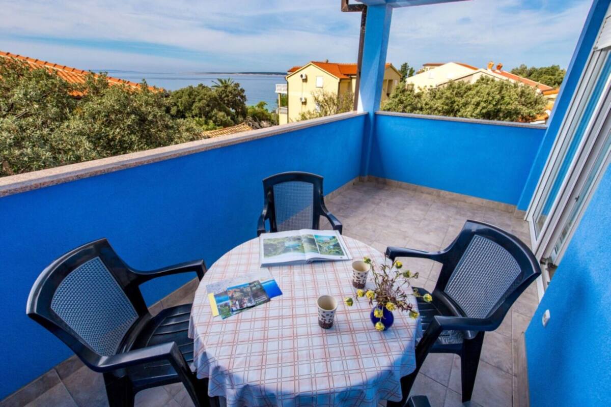Un balcon avec un mur bleu, une table ronde recouverte d’une nappe à carreaux, deux chaises noires et un petit vase avec des fleurs jaunes.