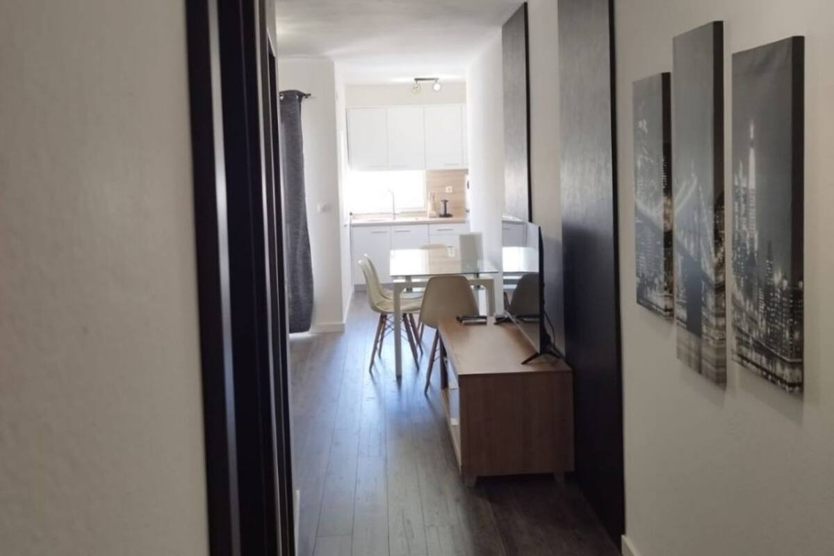 A narrow hallway with a wooden floor, a desk with a computer, and a chair. There are three framed pictures on the wall.