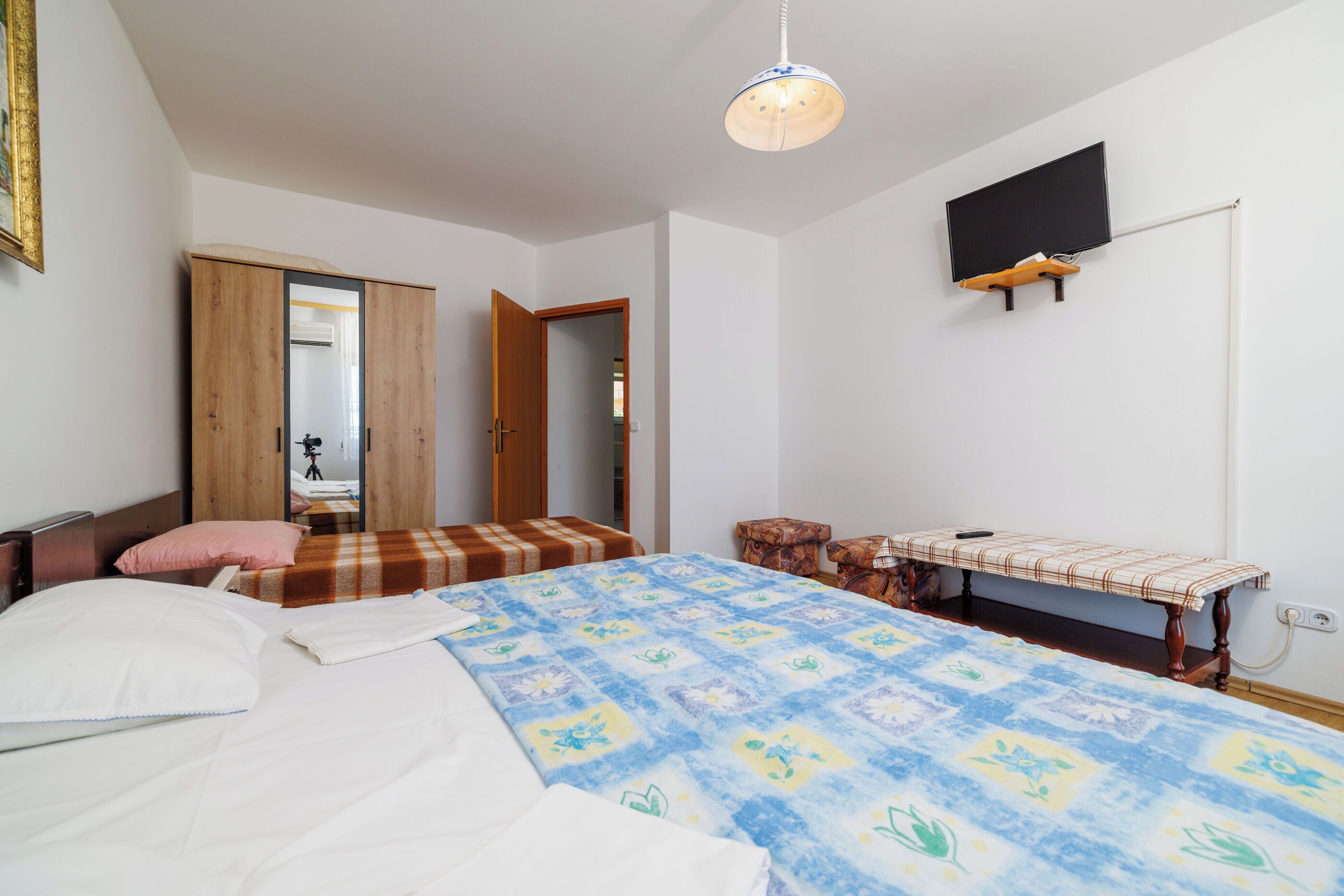A bedroom with a bed, a bench, a television mounted on the wall, and a door leading to another room.