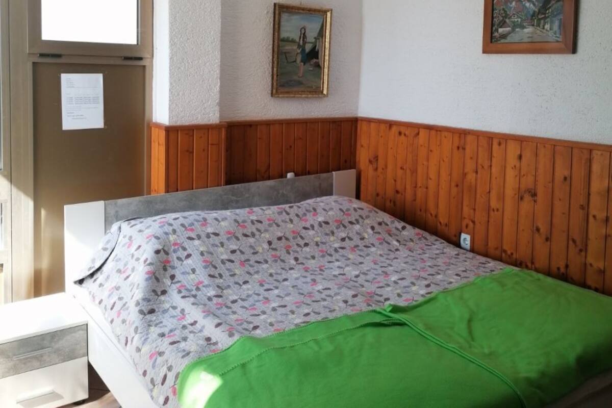 A bedroom with a bed, wooden headboard, two framed pictures, a window, and a door with a bulletin board.