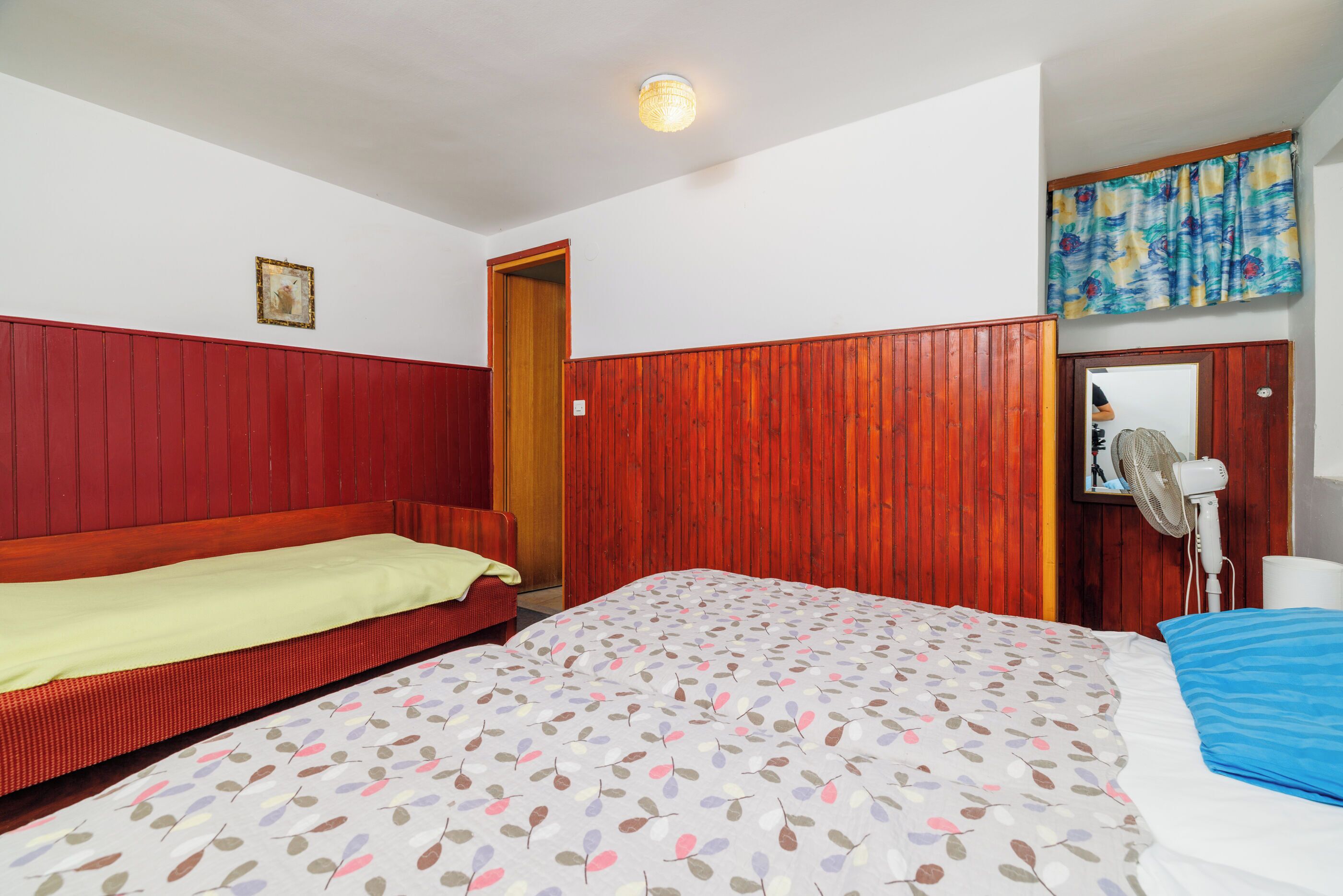 A room with a bed, a wooden wardrobe, a mirror, and a fan.