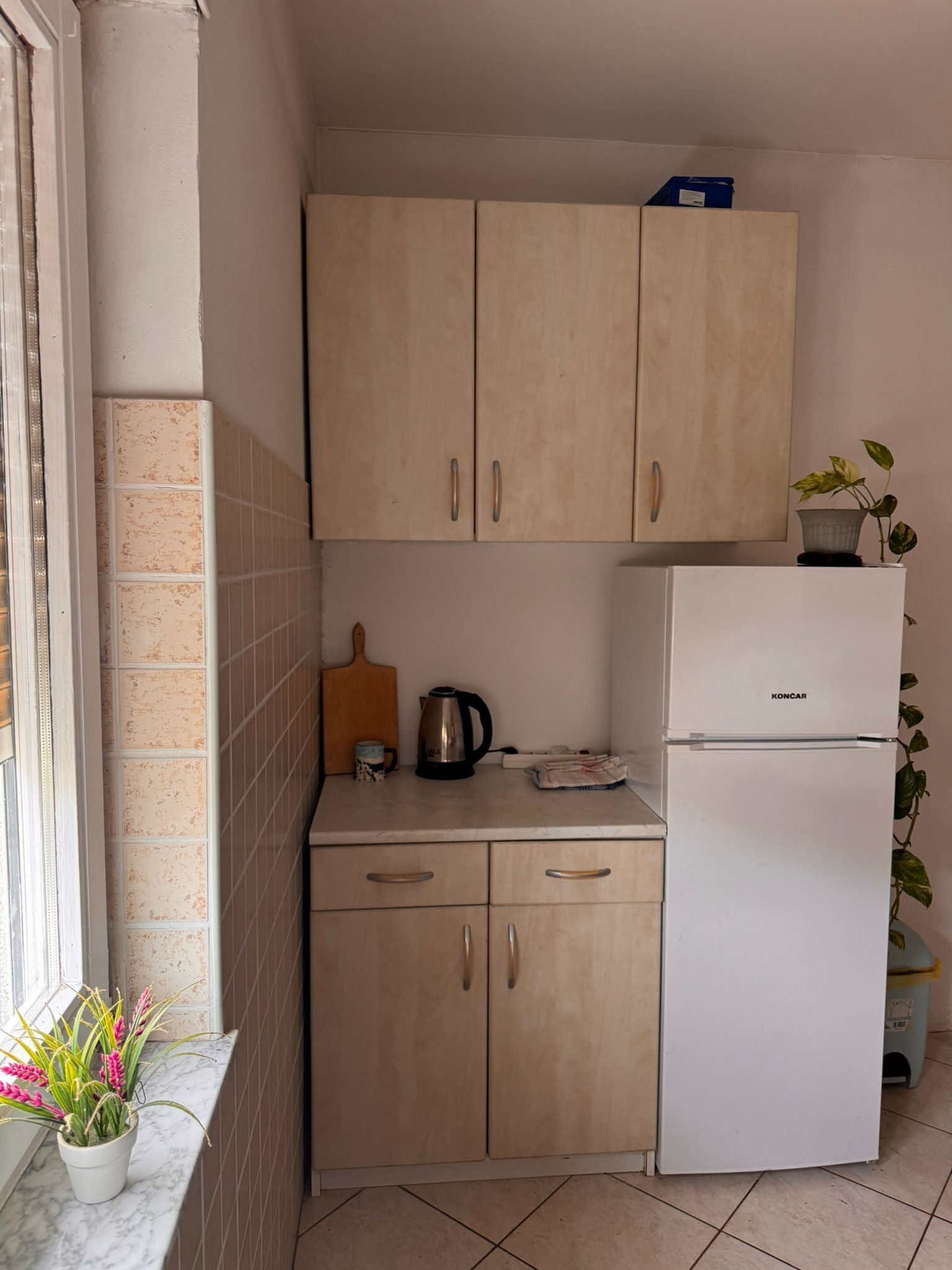 Una cuina senzilla amb armaris beix, un frigorífic blanc i una llosa ceràmica a la paret.