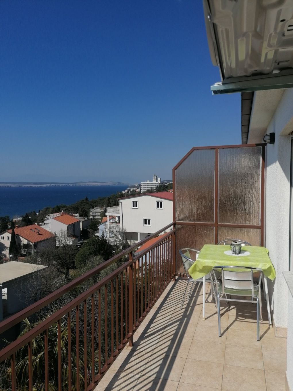Un balcon avec une table et des chaises, donnant sur une ville côtière avec ses bâtiments et la mer.