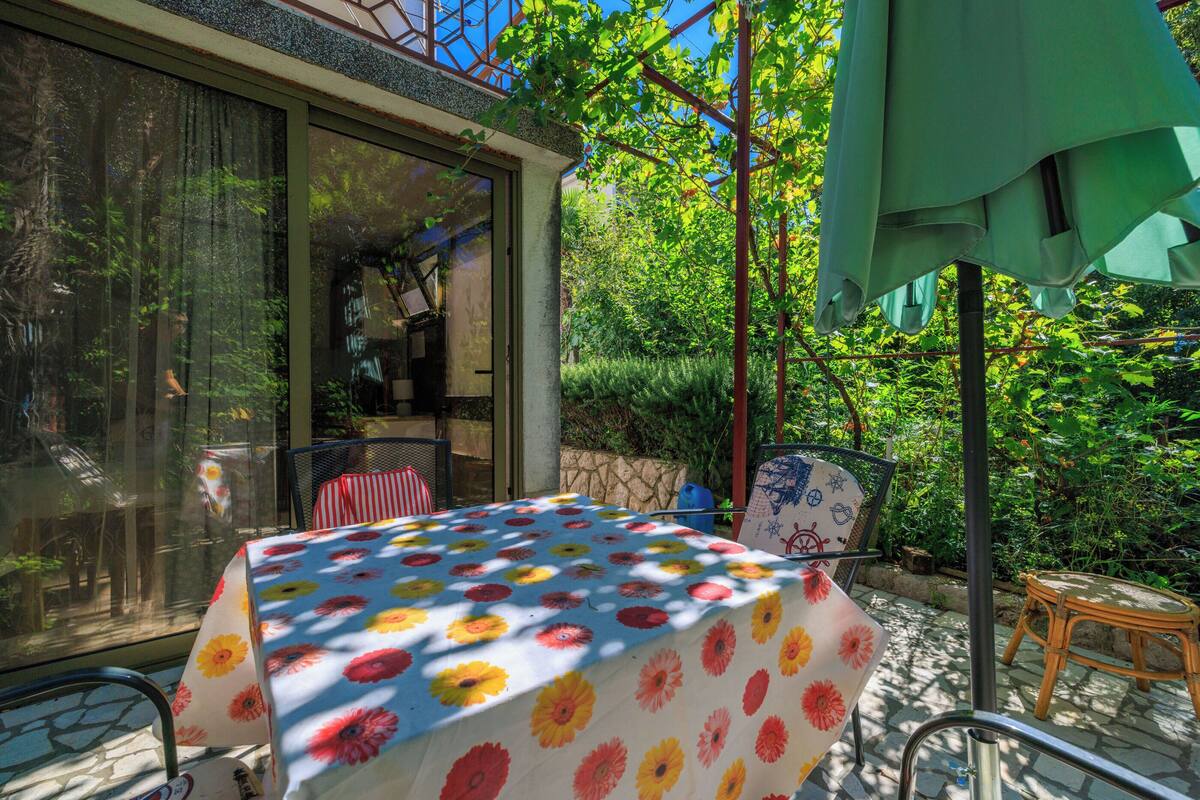 An outdoor patio with a table covered in a floral tablecloth, surrounded by greenery and a glass door leading to a room.