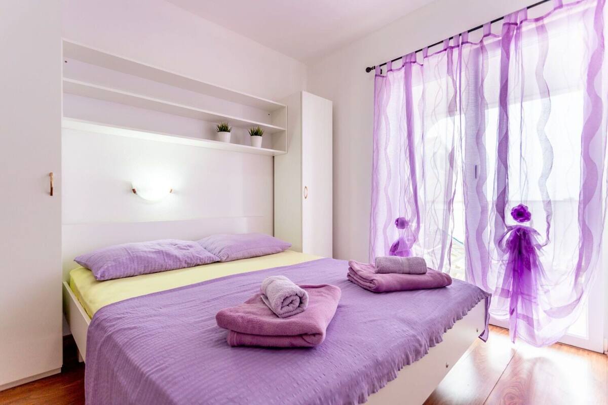 A bedroom with a bed, purple bedding, a window with purple curtains, and a small shelf with plants.