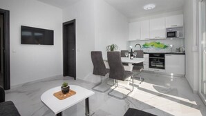 Un espace repas moderne et bien éclairé, avec une table blanche, quatre chaises grises et une petite plante en pot. Une porte sombre et un téléviseur à écran plat sont visibles sur le mur.