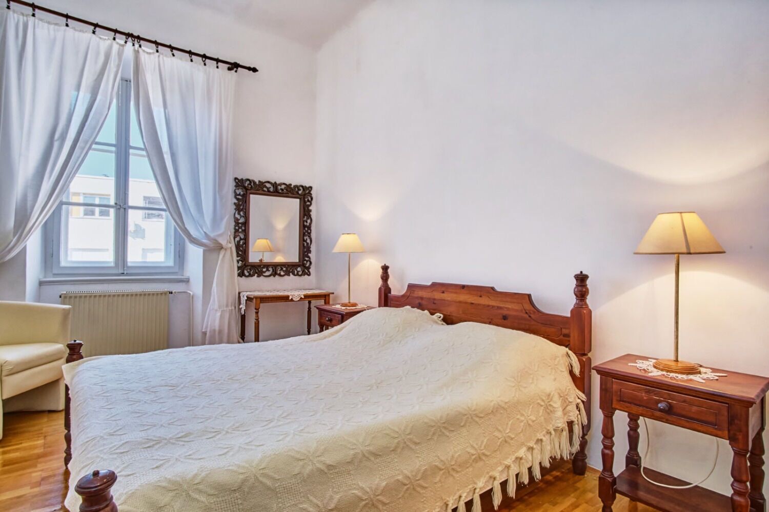 A bedroom with a large bed, two bedside tables with lamps, a wooden headboard, a chair, a window with curtains, and a mirror on the wall.
