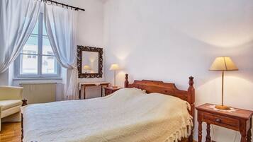 A bedroom with a large bed, two bedside tables with lamps, a wooden headboard, a chair, a window with curtains, and a mirror on the wall.