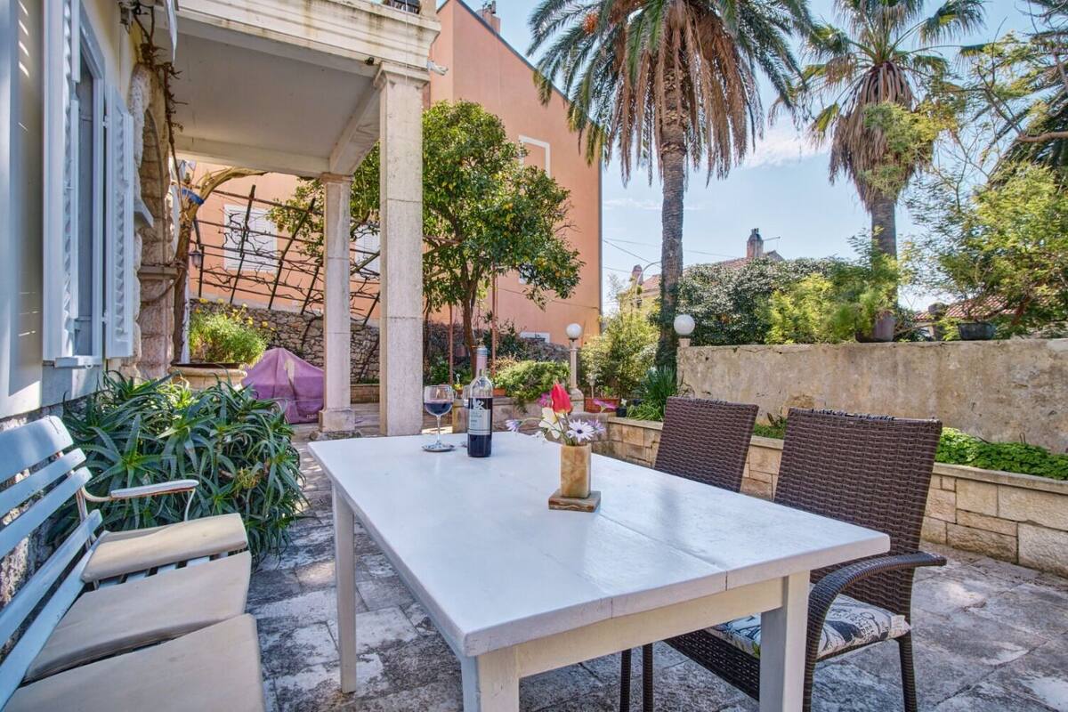 Een terras buiten met een witte tafel, rieten stoelen, een fles wijn en een bloevas.