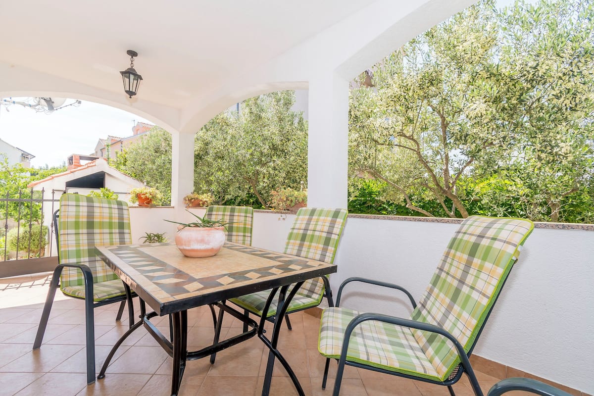 Une terrasse couverte, agrémentée d’une table et de chaises, entourée de verdure et de bâtiments.