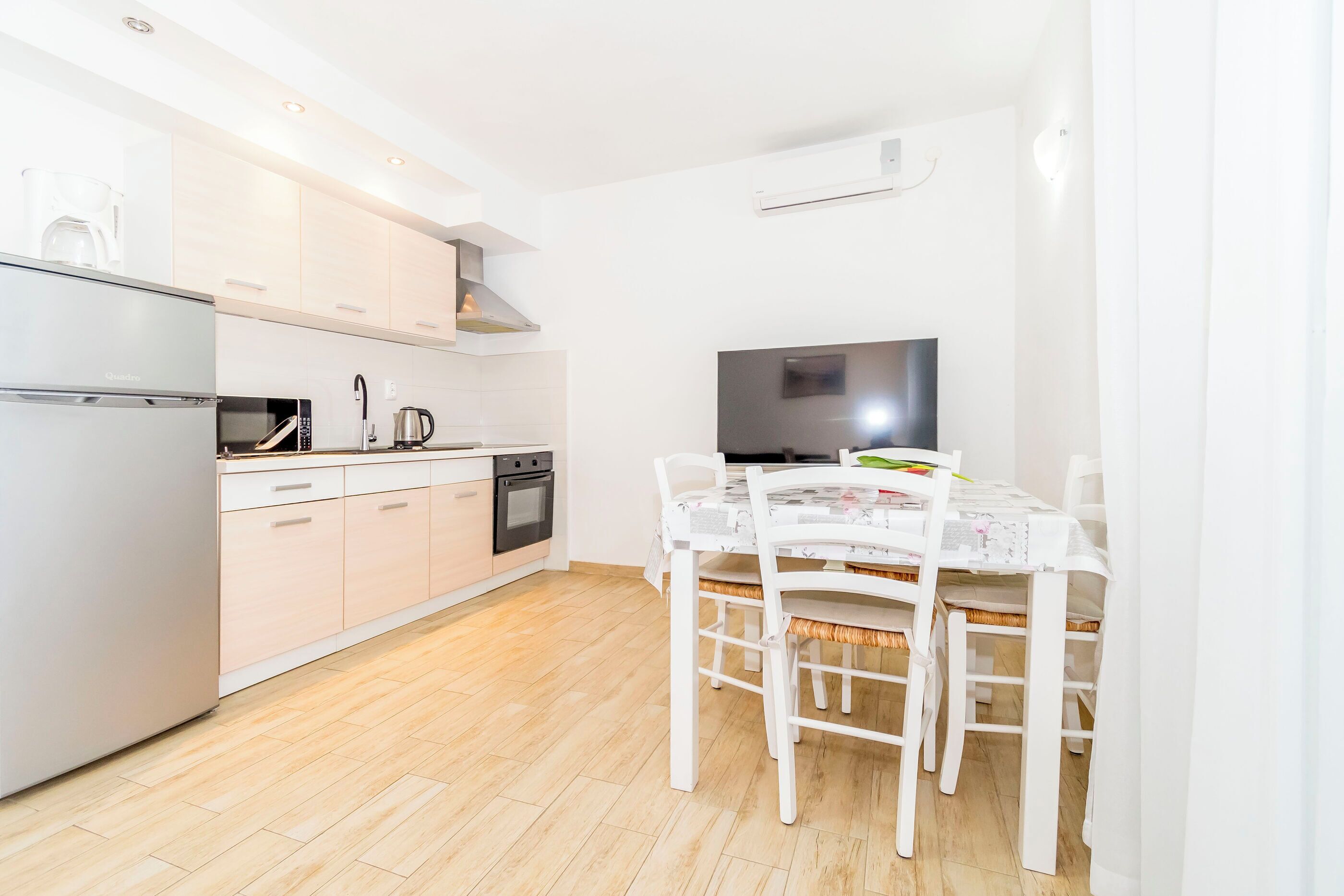 A modern kitchen with a refrigerator, microwave, stove, and sink. There is a dining table with chairs and a flat-screen TV.
