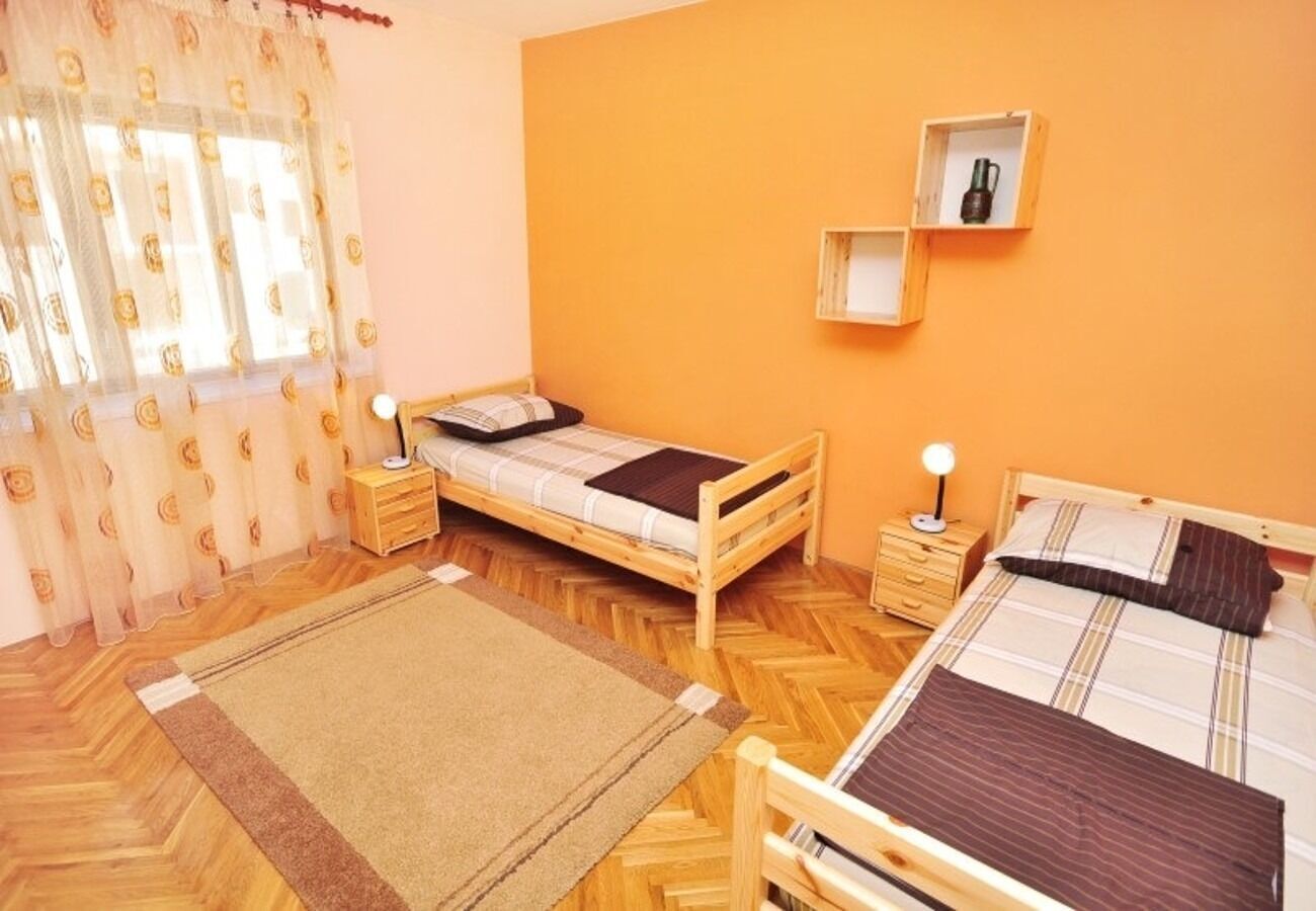 A room with two wooden beds, a small wooden nightstand with a lamp, a shelf with a bottle, and a window with sheer curtains.