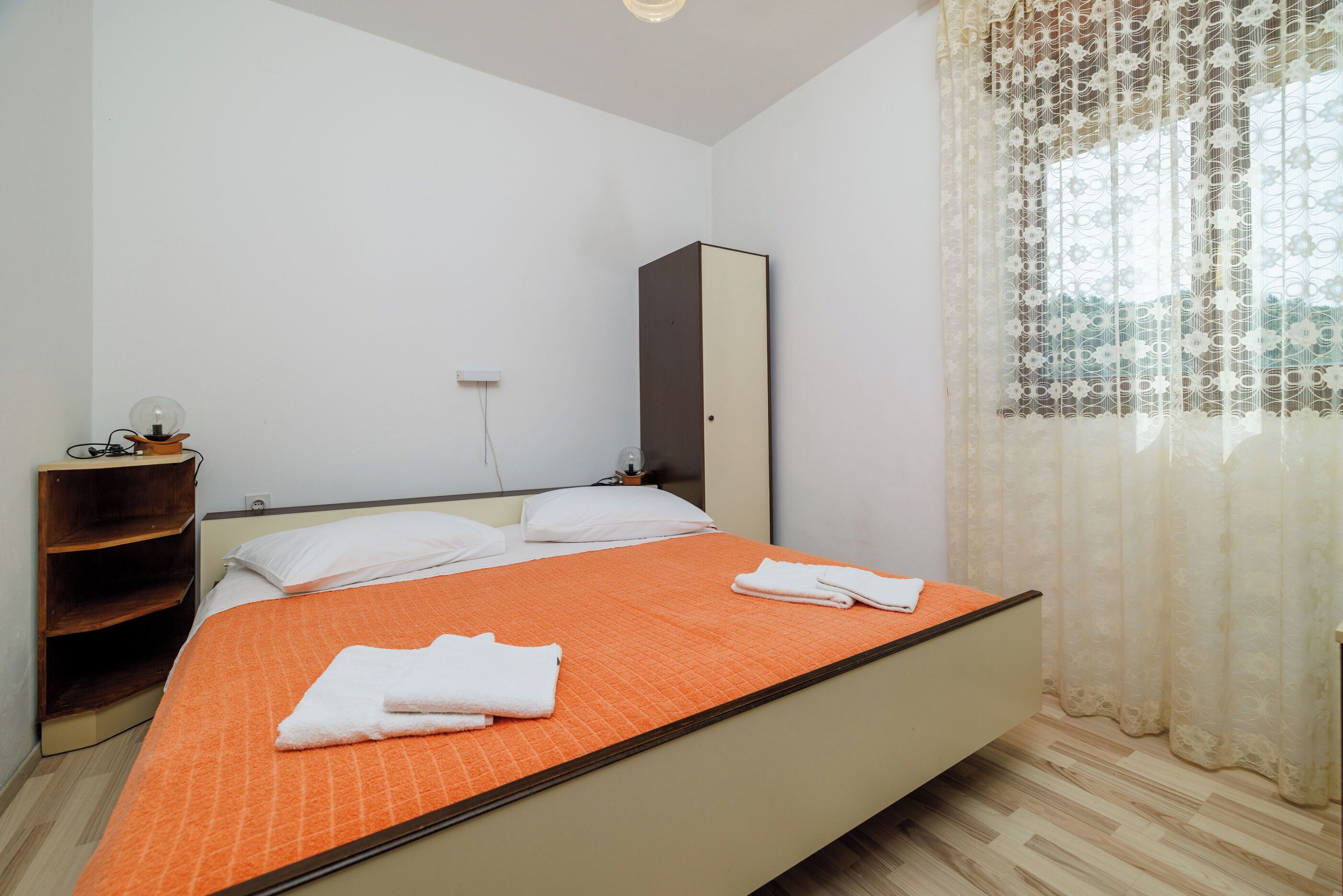 Habitación de hotel con cama, armario, mesita de noche y ventana con cortinas translúcidas.