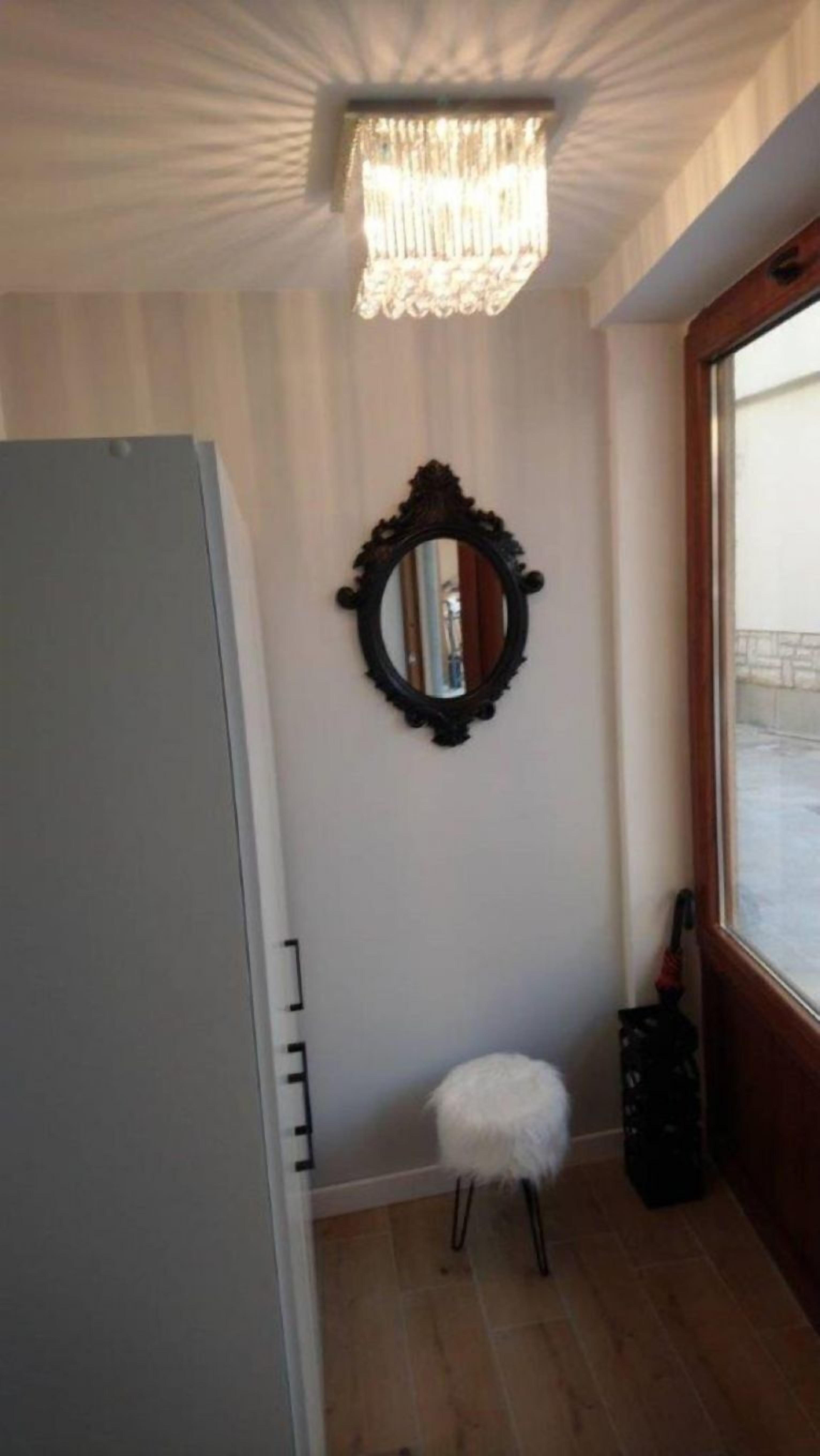 A room with a wooden floor, a mirror on the wall, a small stool, and a window with a view of a building outside.