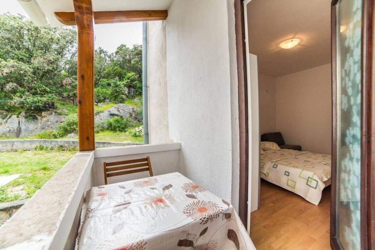 A balcony with a table and chair, a bed with a patterned bedspread, and a view of greenery outside.