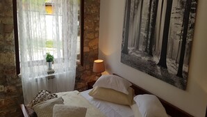 Ein Schlafzimmer mit Bett, Kissen, Nachttisch, Lampe, Fenster mit durchsichtigen Vorhängen, einem Blumentopf und einem Wandbild zum Thema Wald.
