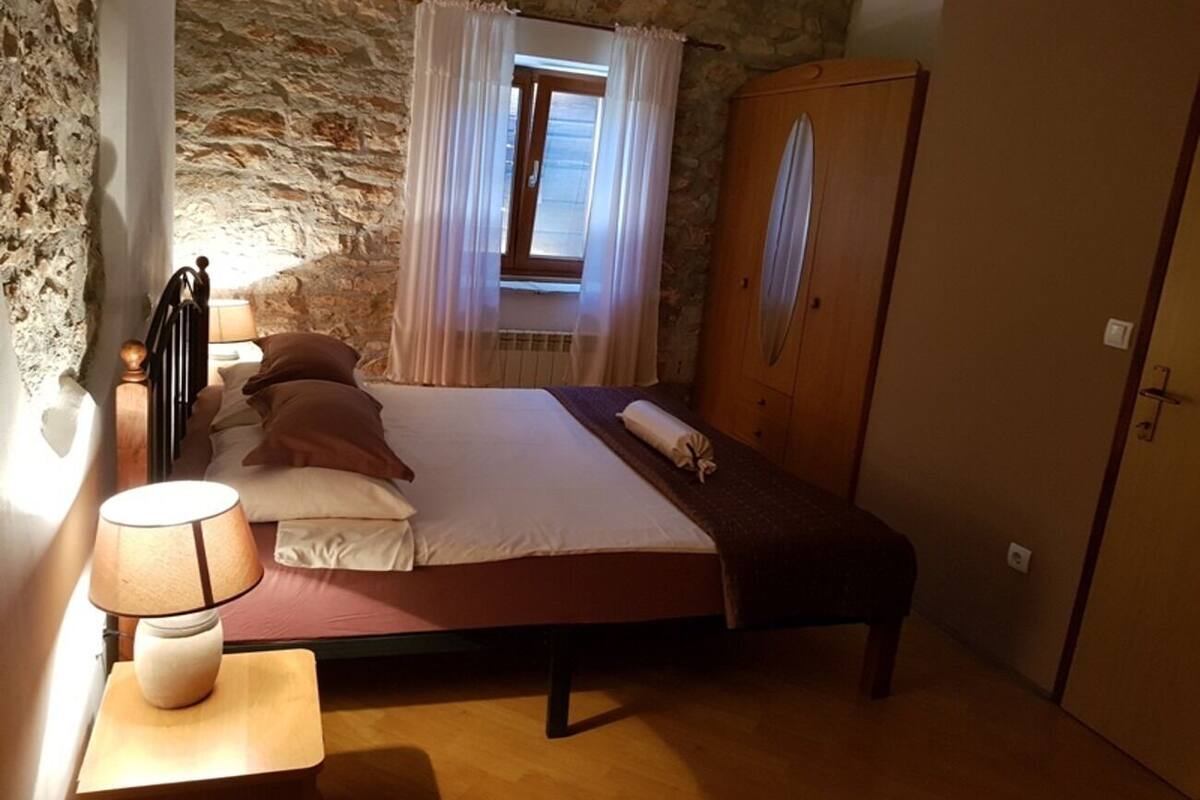 A bedroom with a bed, a bedside table with a lamp, a window with curtains, and a wardrobe.