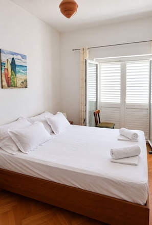 A hotel room with a large bed, a painting of surfboards, a window with blinds, and a wooden floor. - Apartment Anka (Omis)