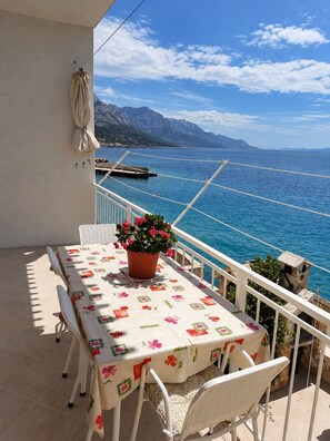 Ein Balkon mit Tisch und Stühlen, einem Blumentopf und Blick auf Meer und Berge.