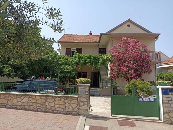 Um edifício de dois andares com fachada de pedra, telhado de telhas vermelhas e um portão verde com a inscrição "APARTMANI".