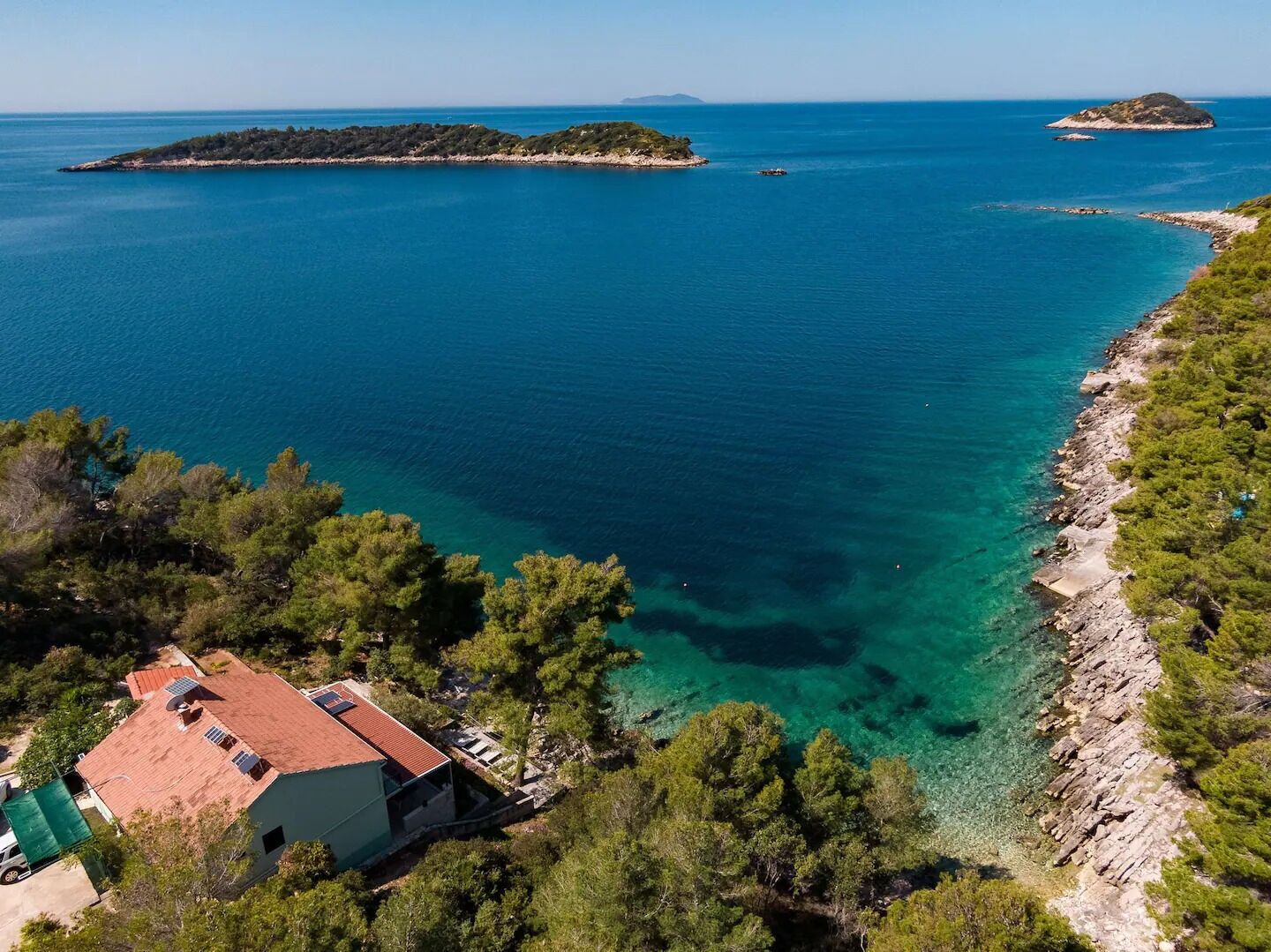 En fastighet vid kusten med en byggnad som har ett rött tak, kristallklart blått vatten och en stenig strand.