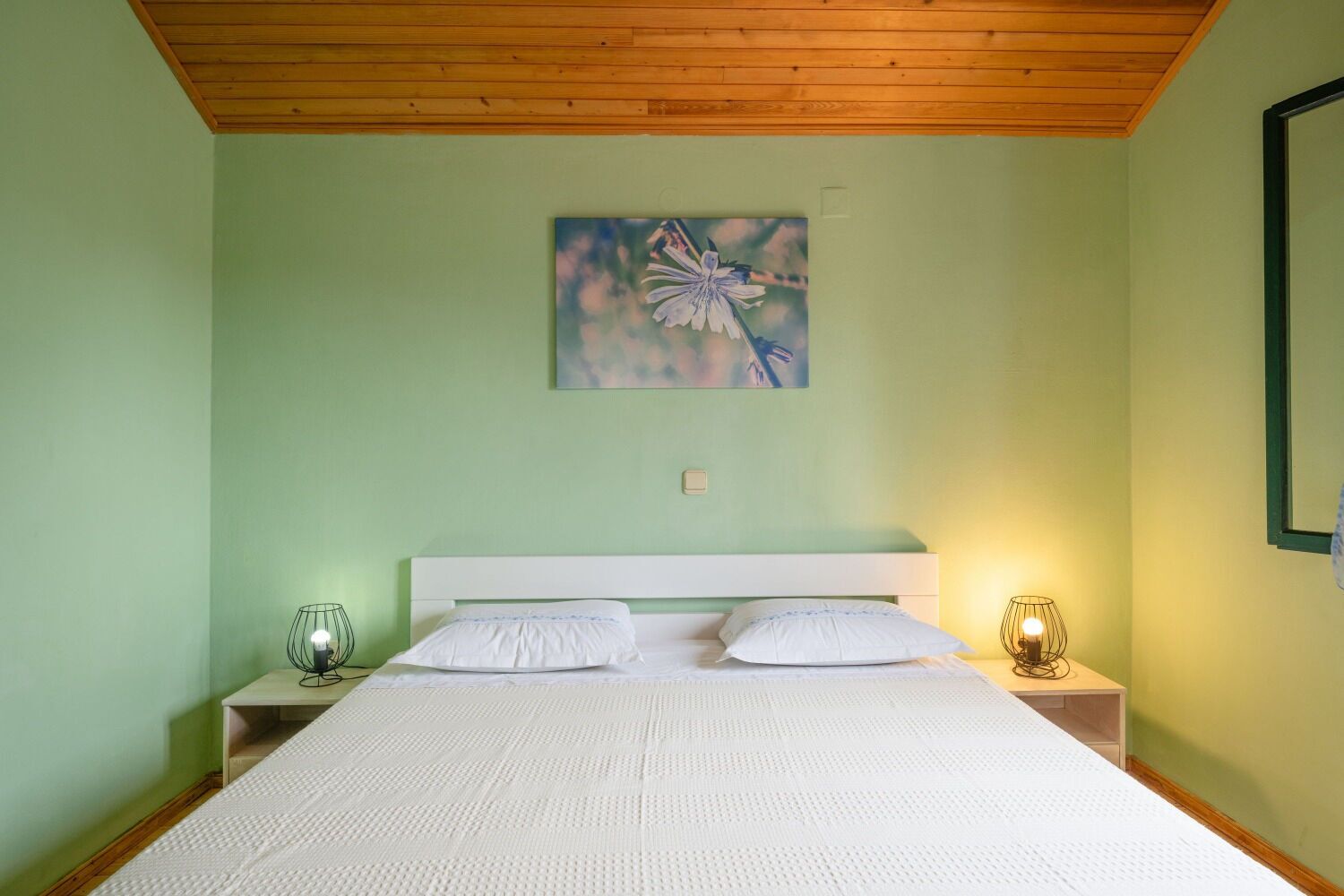 A bedroom with a bed, two bedside lamps, a painting on the wall, and a wooden ceiling.