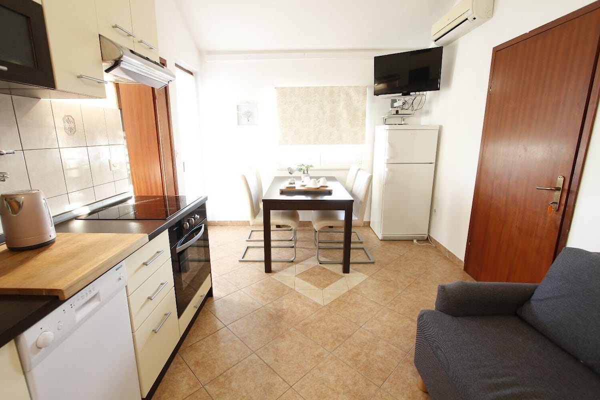 A compact kitchen with a stove, oven, and dishwasher, a dining table with chairs, a TV mounted above a white refrigerator, and a wooden door.
