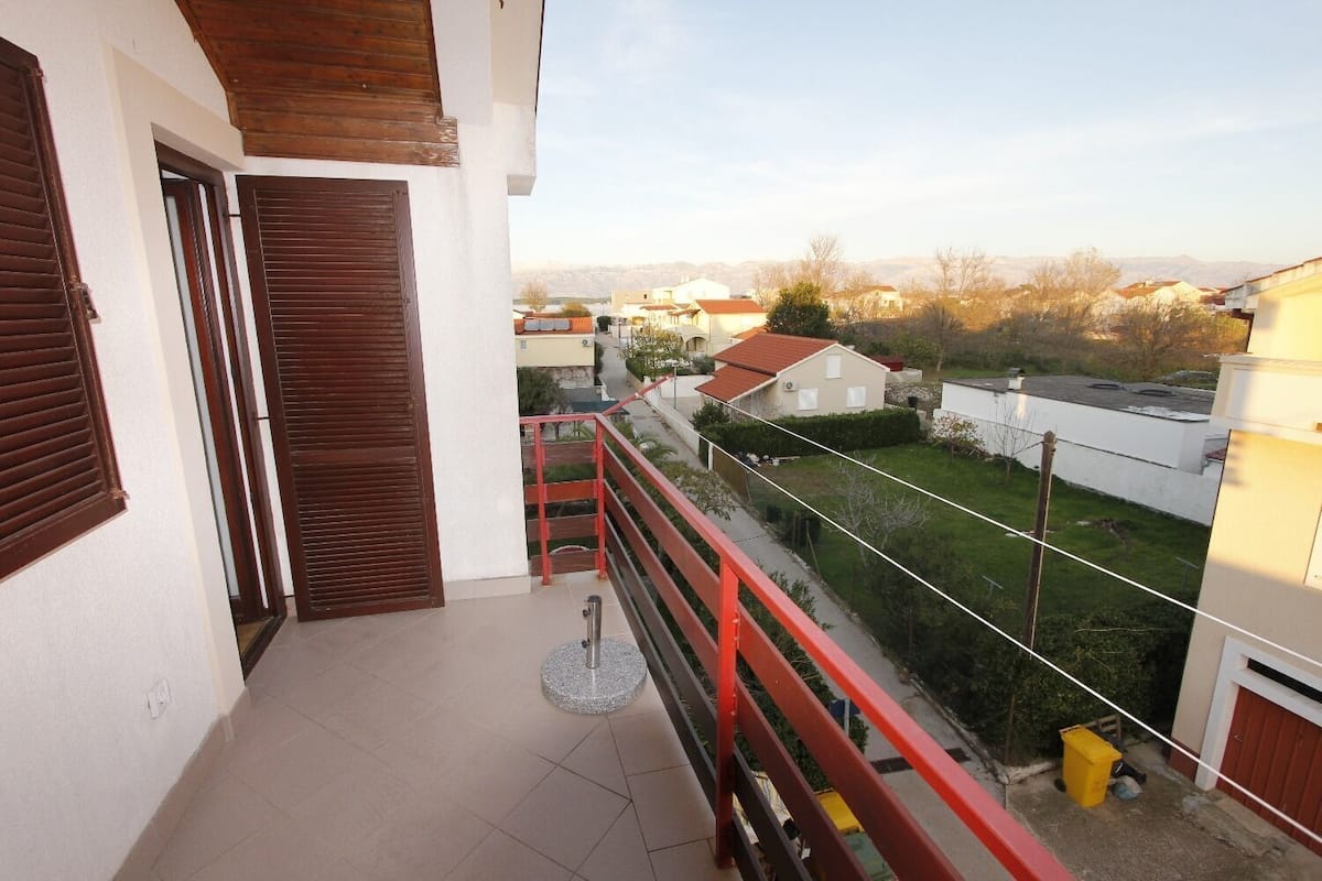 A balcony with a red railing, a wooden door, and a view of neighboring houses and a grassy area.