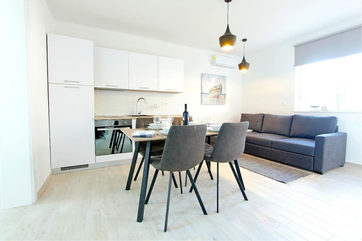 A modern kitchen with white cabinets, a dining table with chairs, and a gray sofa in a living area.