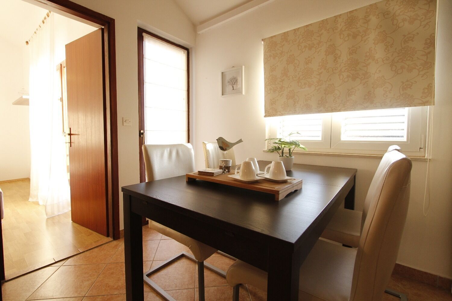 A dining area with a dark wooden table, white chairs, a tray with cups and a bird sculpture, and a window with blinds.