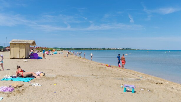 Una playa con gente tomando el sol y nadando, una torre de salvavidas y un cielo azul despejado.