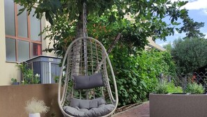 Una silla colgante con cojín y almohada, ubicada en un patio de ladrillo con jardín al fondo.