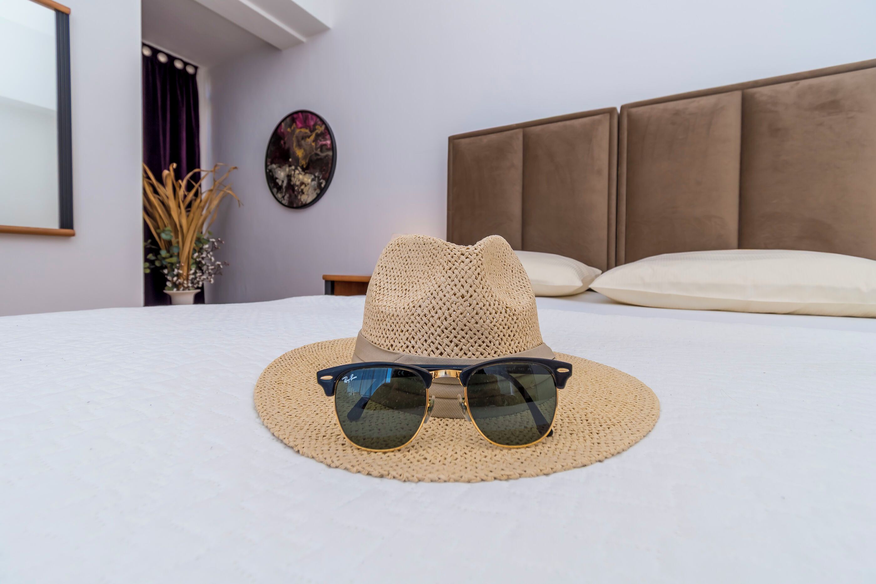 Una cama con un sombrero tejido y gafas de sol encima, un cabecero, una obra de arte en la pared y un jarrón con plantas secas.