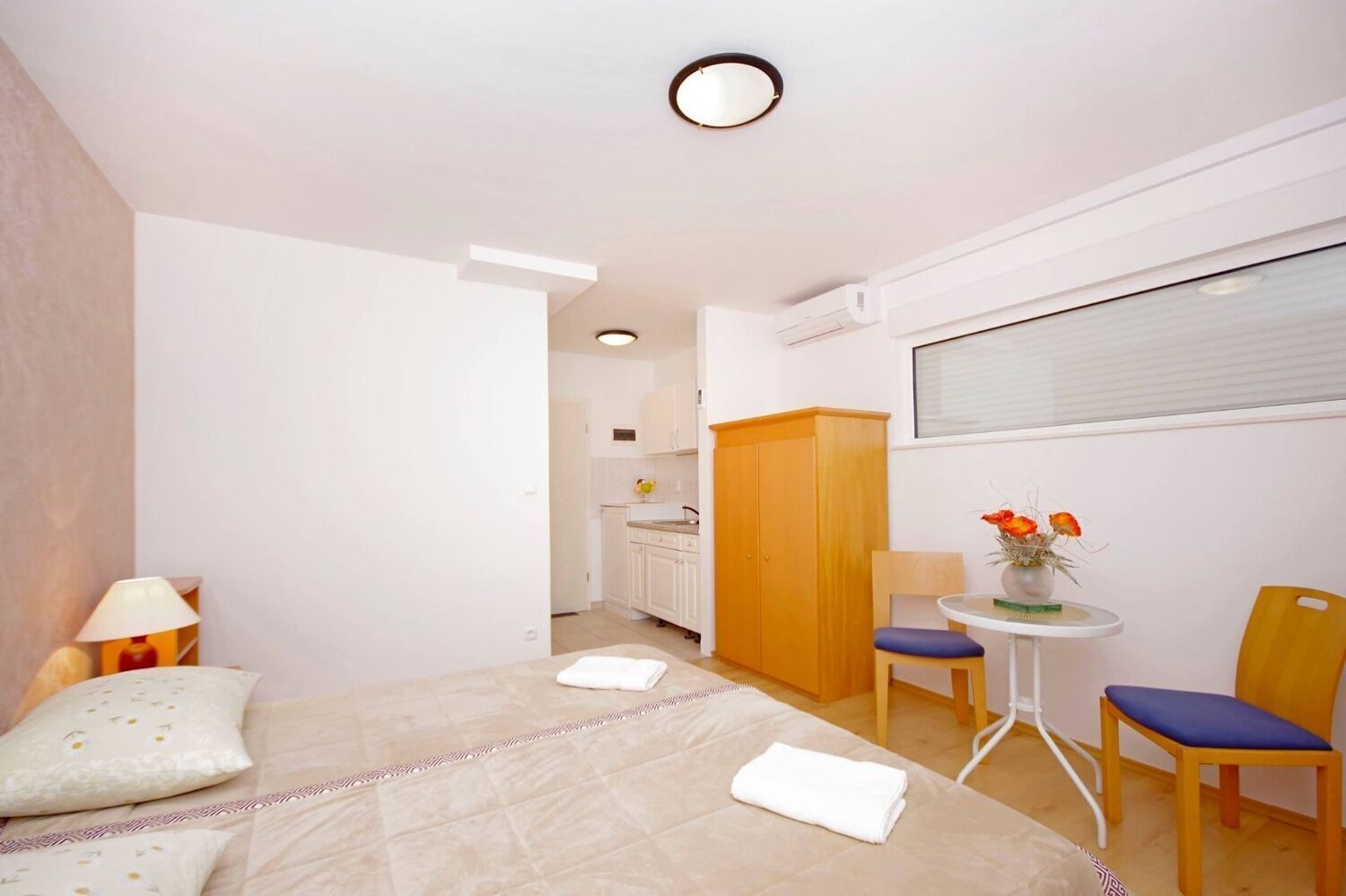 A bedroom with a bed, two chairs, a small table with a vase of flowers, and a window with blinds.