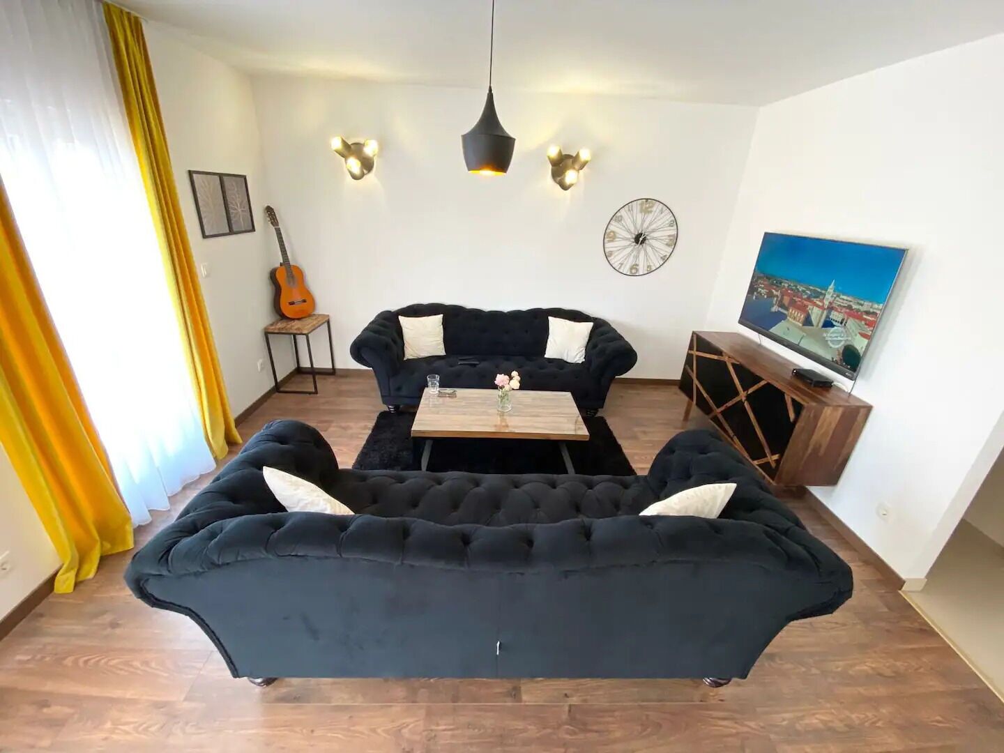 Un salon avec un canapé noir, une table basse en bois, une guitare et un téléviseur posé sur un meuble en bois.
