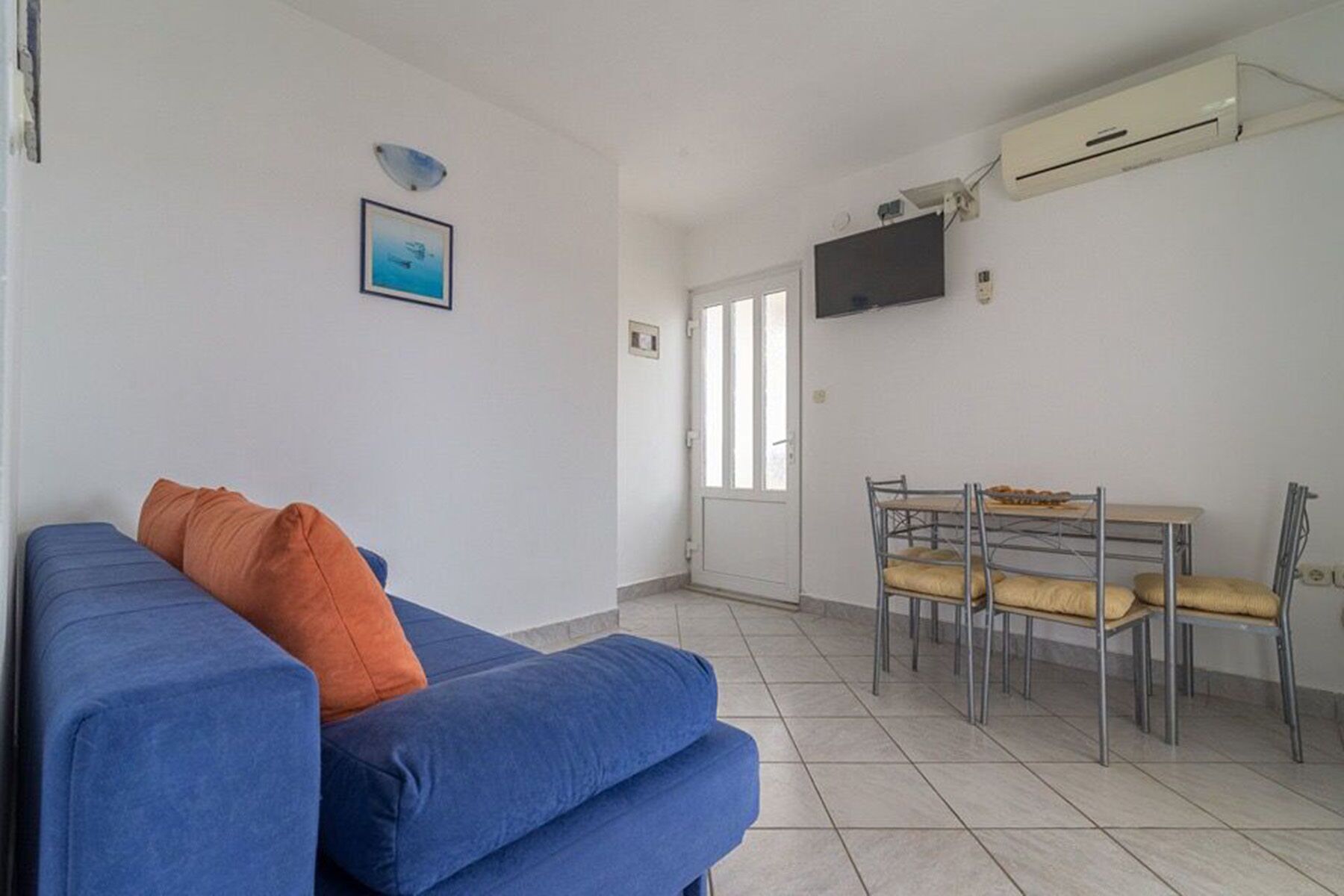 A room with a blue sofa, an orange pillow, a dining table with chairs, a TV mounted on the wall, and an air conditioning unit.