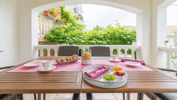 A dining table with a pink placemat, white plates, a glass jar, and a tray with fruits and pastries. - Apartments Sandra (Obrovac)