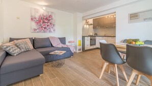 A modern living room with a sofa, a dining table with chairs, a kitchen in the background, and a floral painting on the wall. - Apartments Sandra (Obrovac)